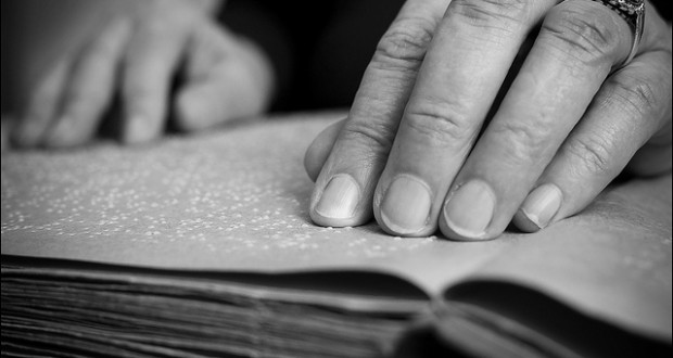 Primer plano en blanco y negro de manos leyendo un libro en braille, con dedos tocando los puntos en relieve sobre la página.